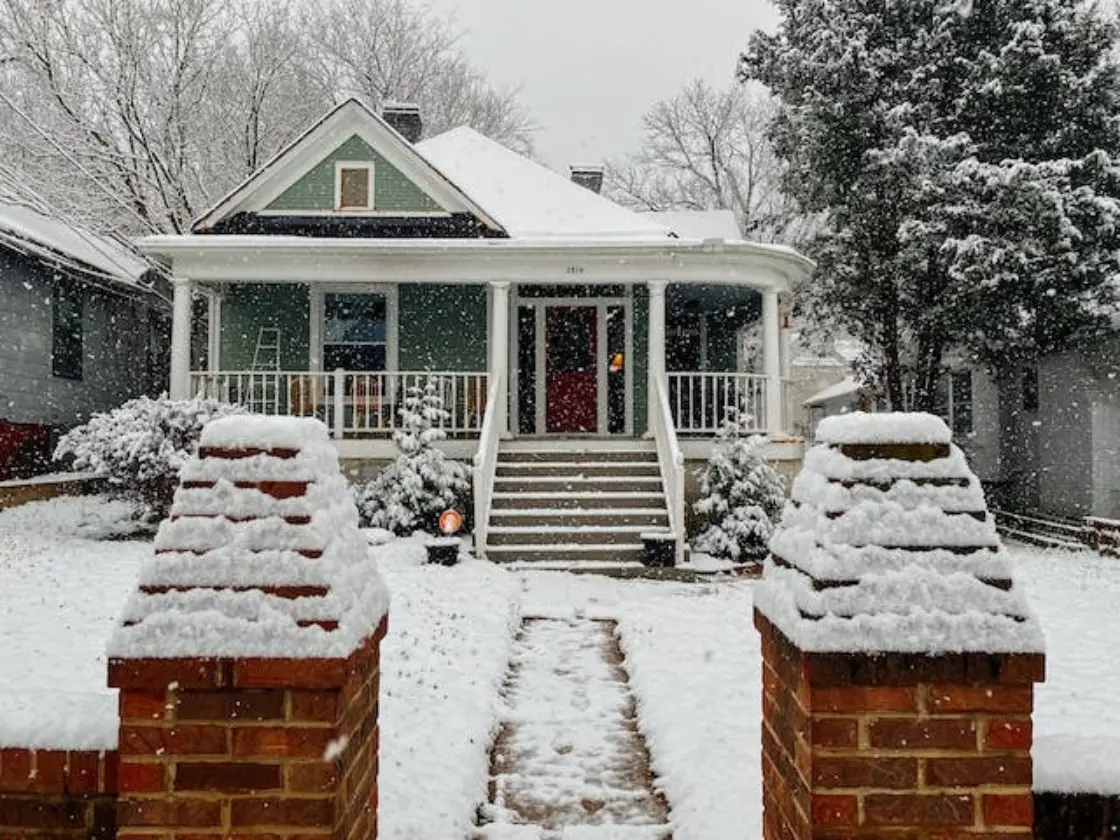 Home with snow on roof, trees, lawn, and sidewalk