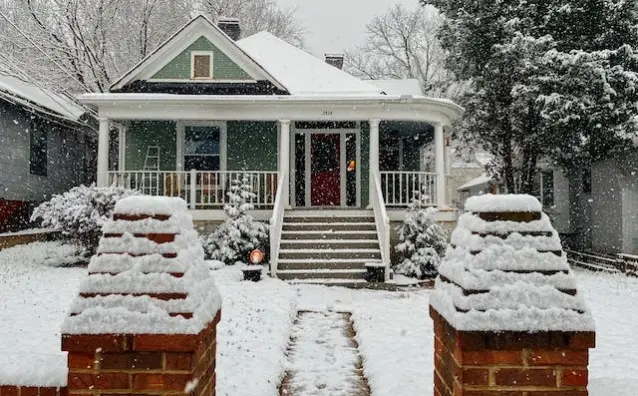 Home with snow on roof, trees, lawn, and sidewalk