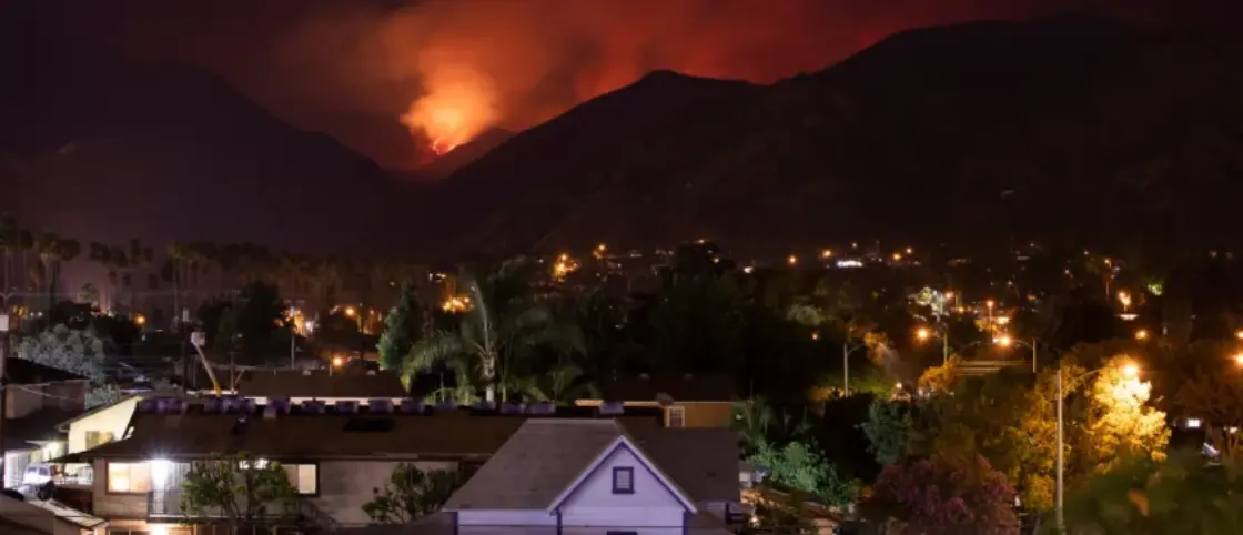 Nighttime and wildfire in background behind mountains approaching neighborhood in foreground.