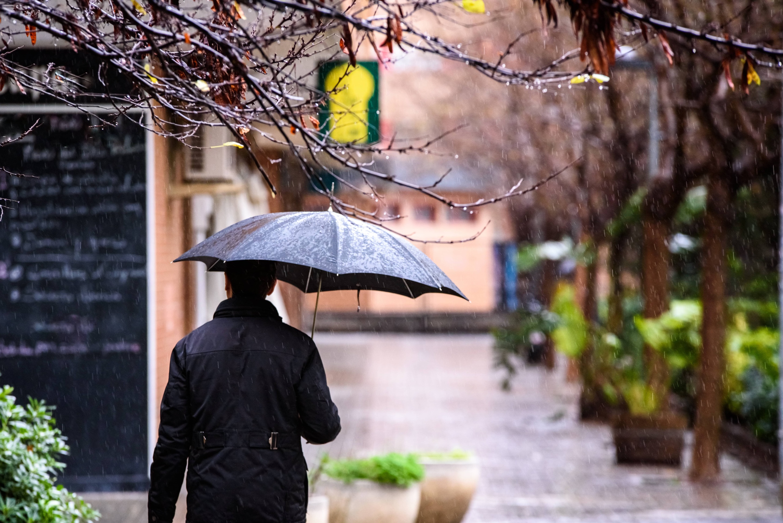 An adult man walked down the street of a city with a black umbrella on a rainy day.