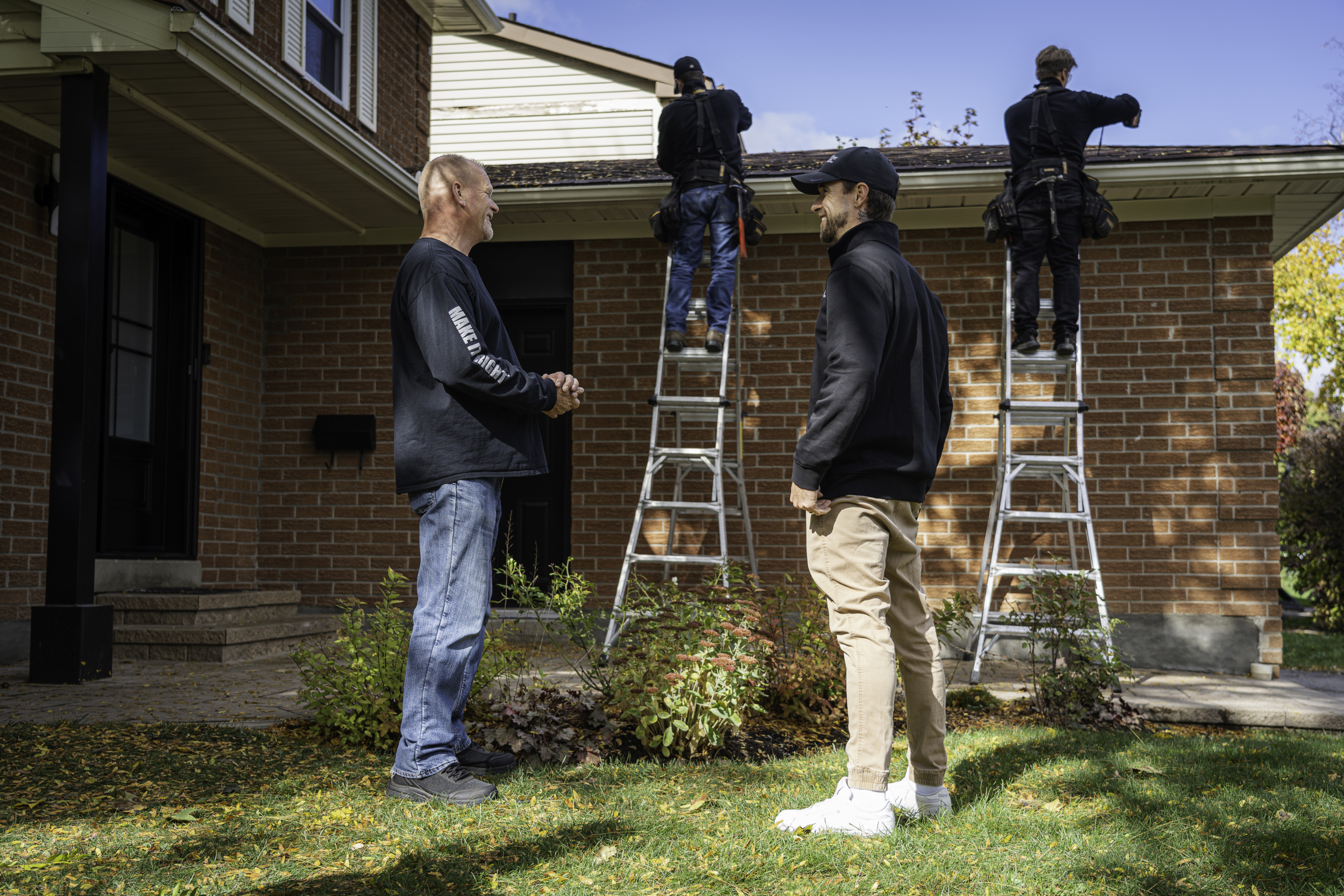 Mike holmes and LeafFilter installer talking in front yard. Background two LeafFilter installers on ladders installing LeafFilter gutter guards on brick home.