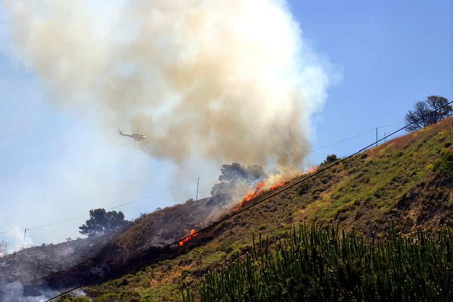 hillside with vegetation on fire and smoke rising helicoptor in background dumping water to fight wildfire awareness