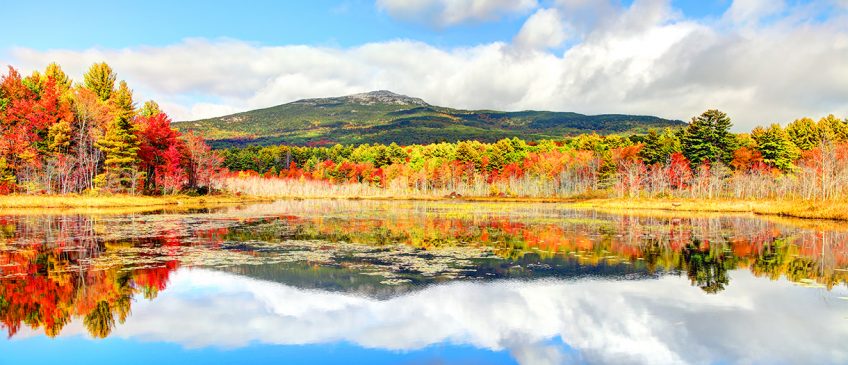 A mountainous New England landscape with a lake in the foreground. The foliage is in hues of gold and red.