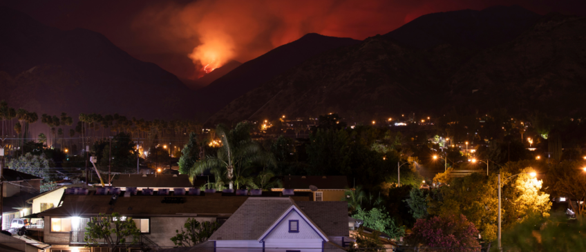 Nighttime and wildfire in background behind mountains approaching neighborhood in foreground.